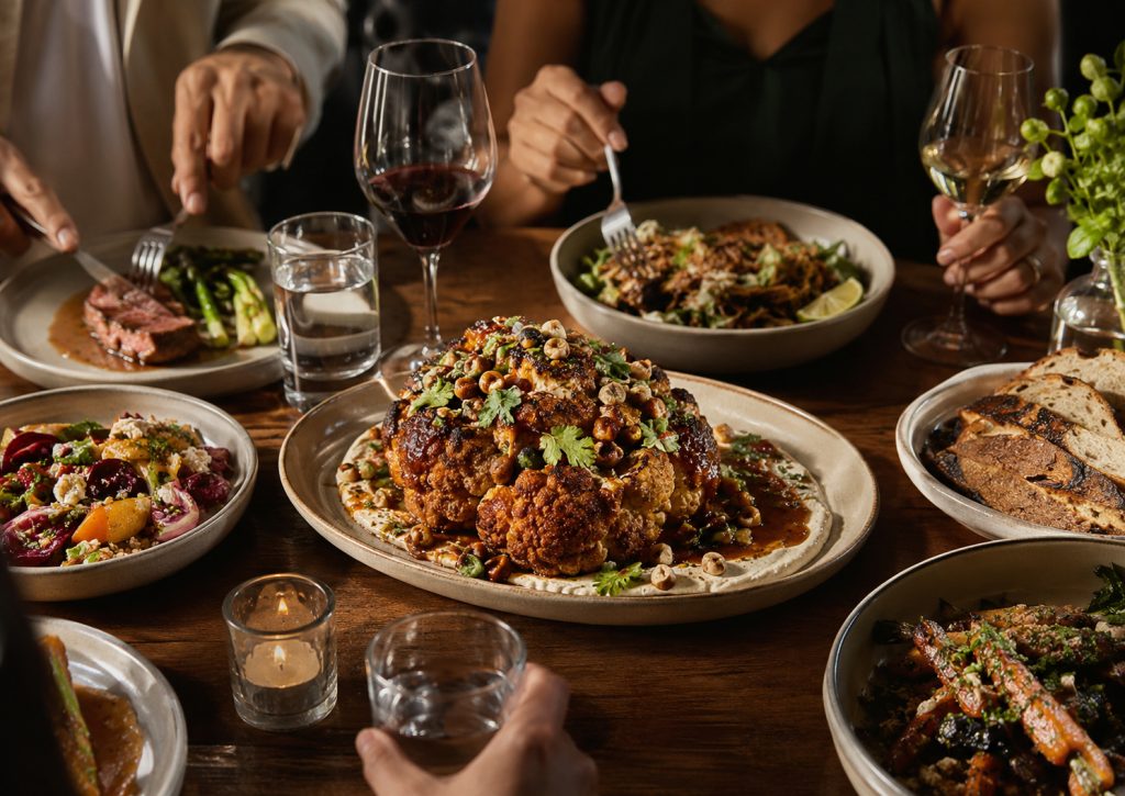 Tavolo di ristorante con piatti condivisi e cavolfiore arrosto al centro, simbolo del plant-based desiderabile.